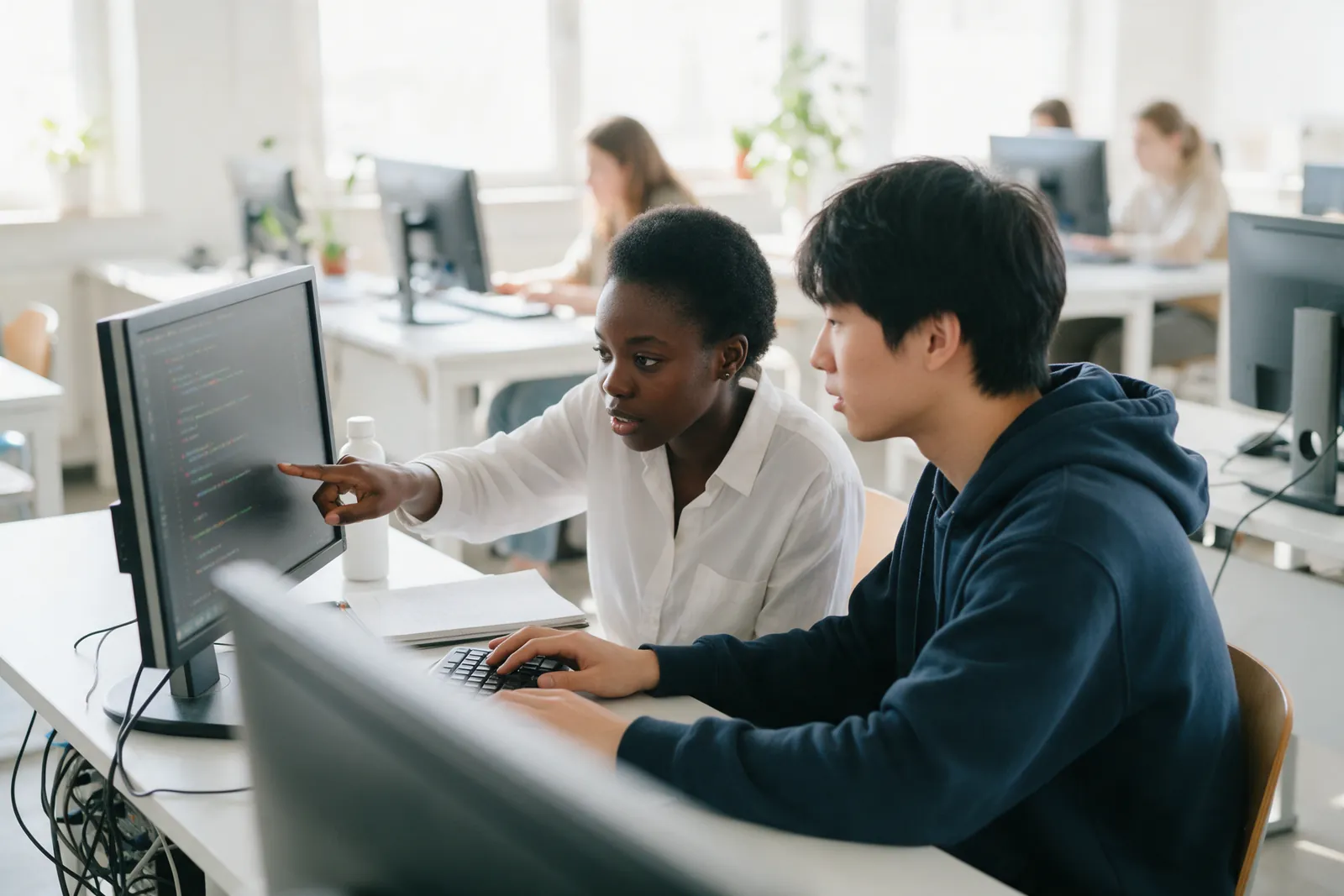 Two students coding side by side at one monitor in a bright computer lab, one pointing at the screen