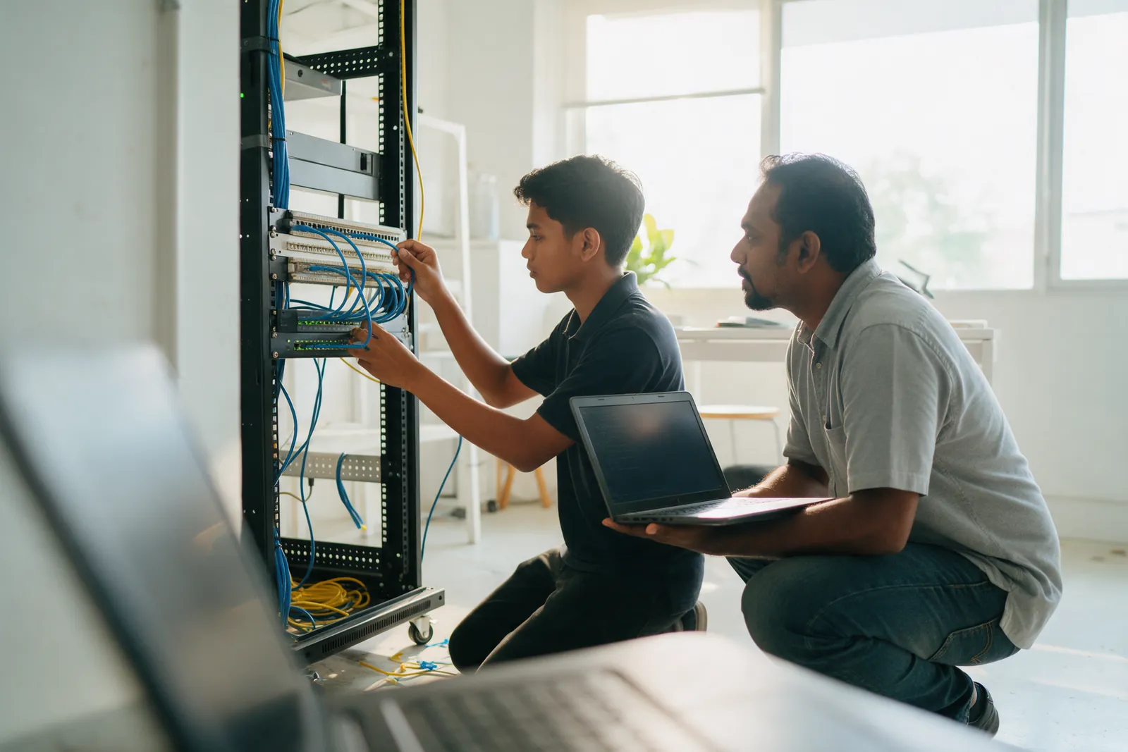 A trainee plugging a patch cable into a network rack while a senior engineer holds a laptop
