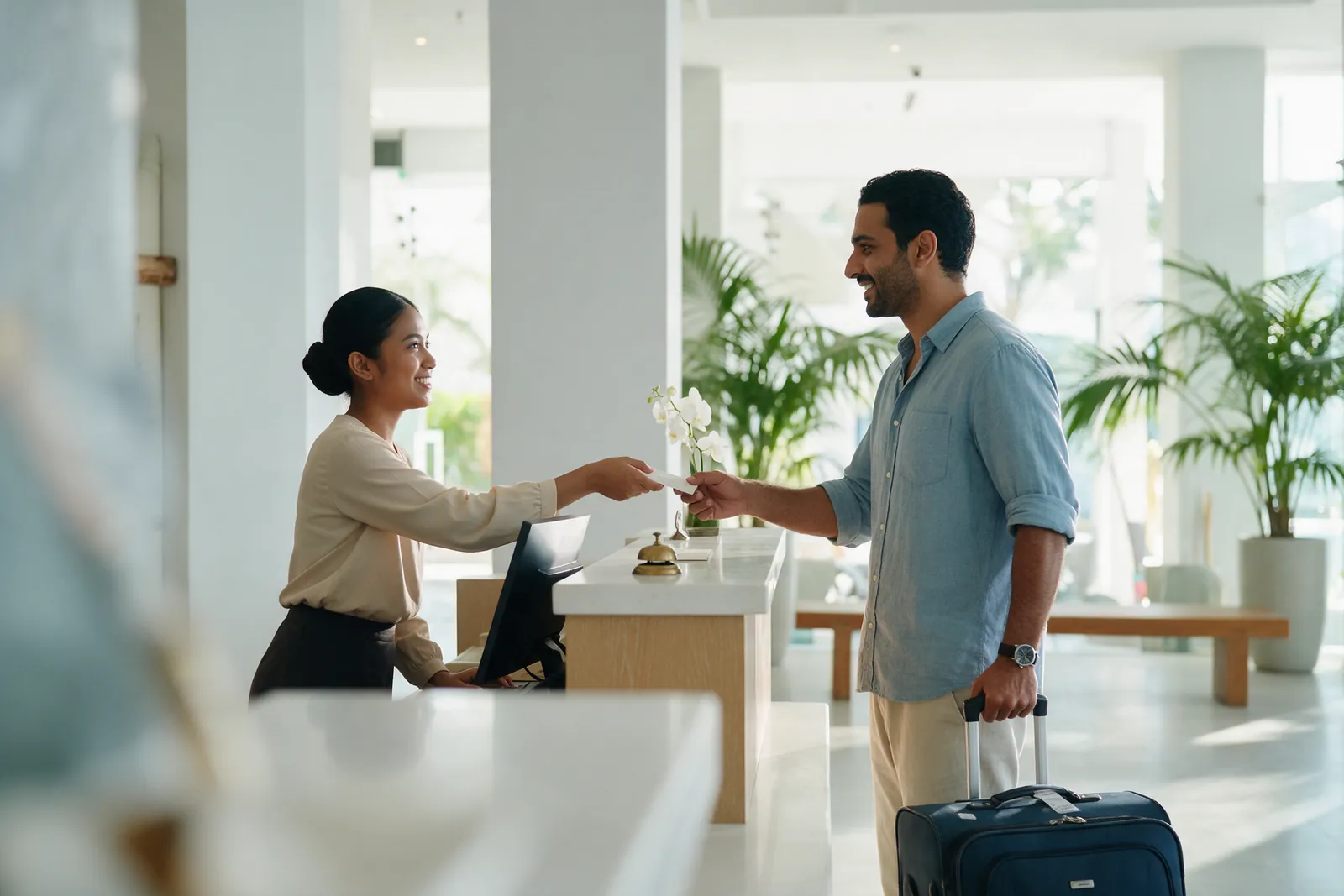 A trainee at a bright wooden hotel front desk handing a key-card folder to a guest, palms behind