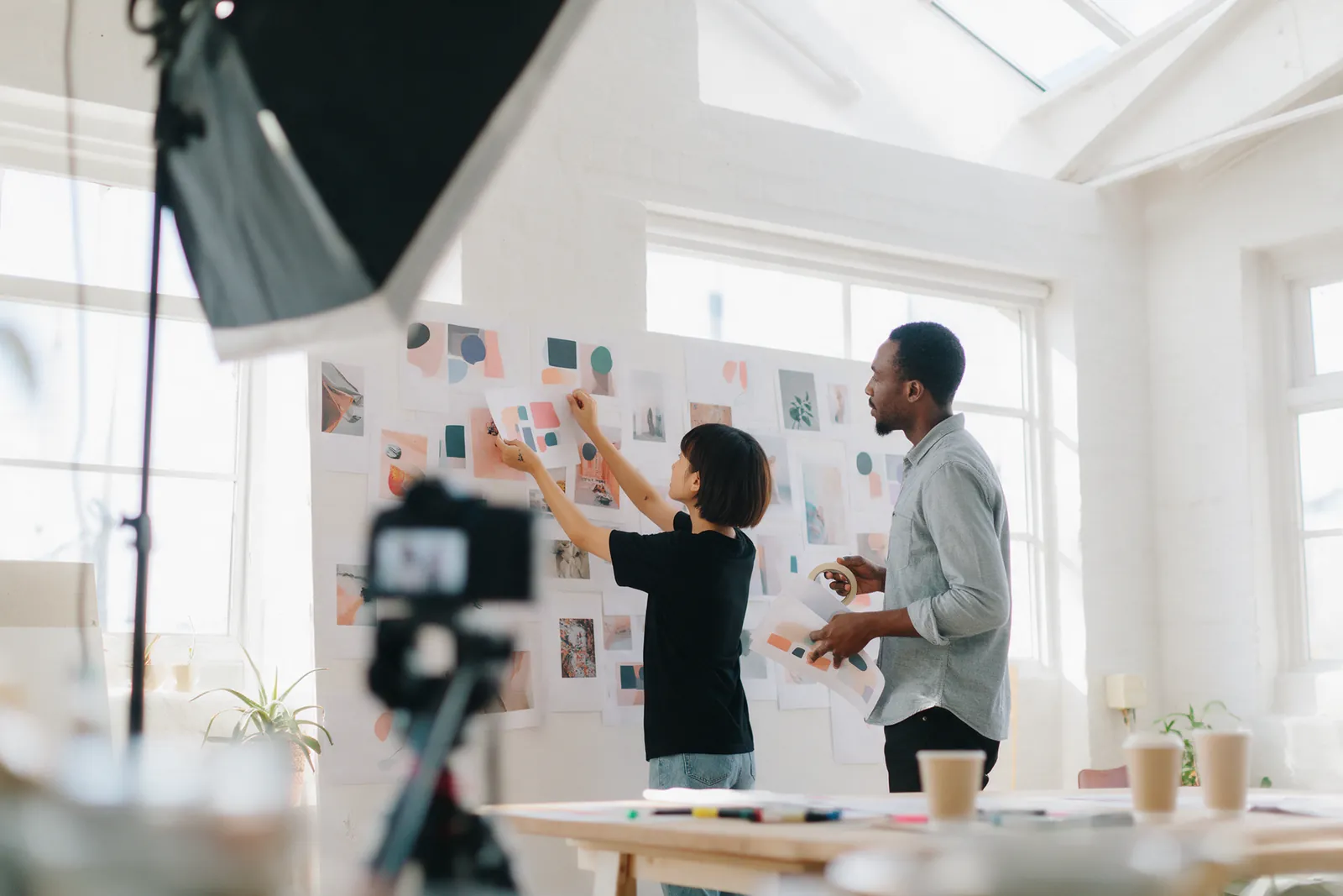 Two people pinning printed layouts to a studio wall, a camera on a tripod in the foreground