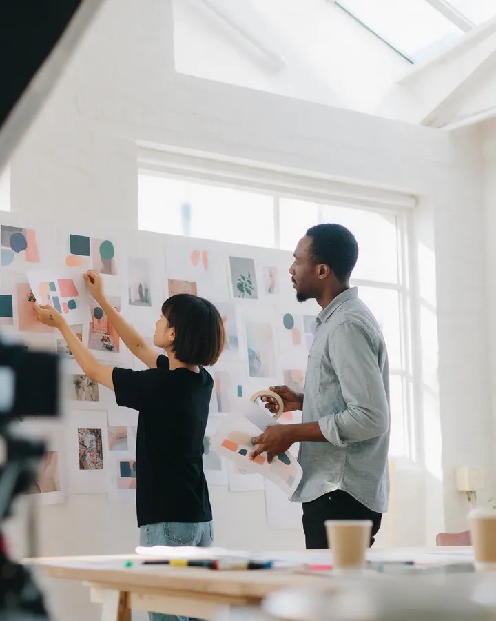 Two people pinning printed layouts to a studio wall, a camera on a tripod in the foreground