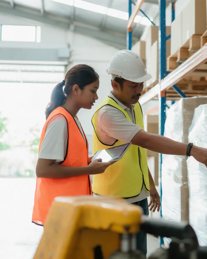 A trainee in a hi-vis vest holding a tablet while a supervisor points down a warehouse aisle