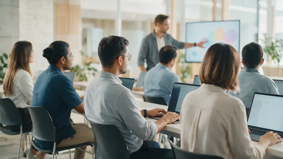 Working adults at a row of laptops while a trainer gestures towards a large screen in a bright training room