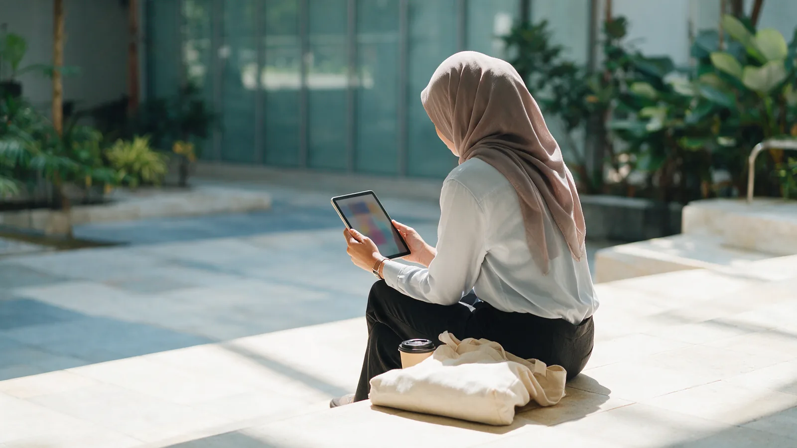 Illustrative photograph: a student sitting on a sunlit stone step in a college courtyard, reading on a tablet