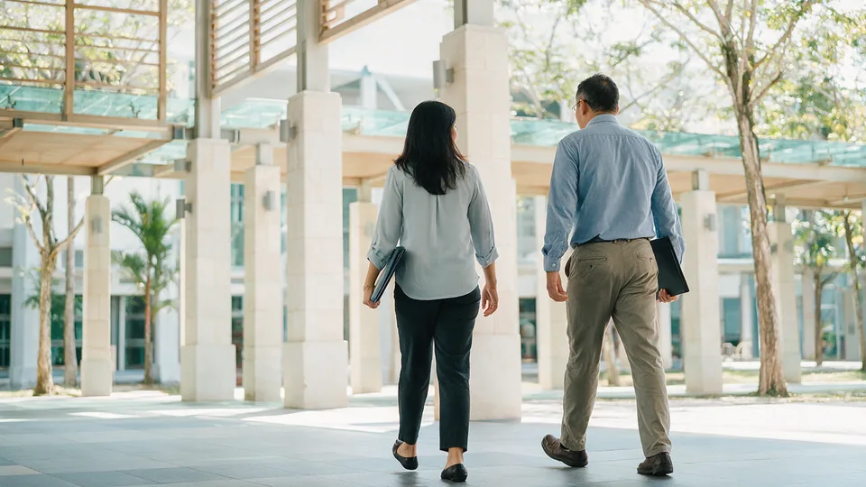 Two academics with folders walking along a sunlit covered walkway on a modern campus, seen from behind