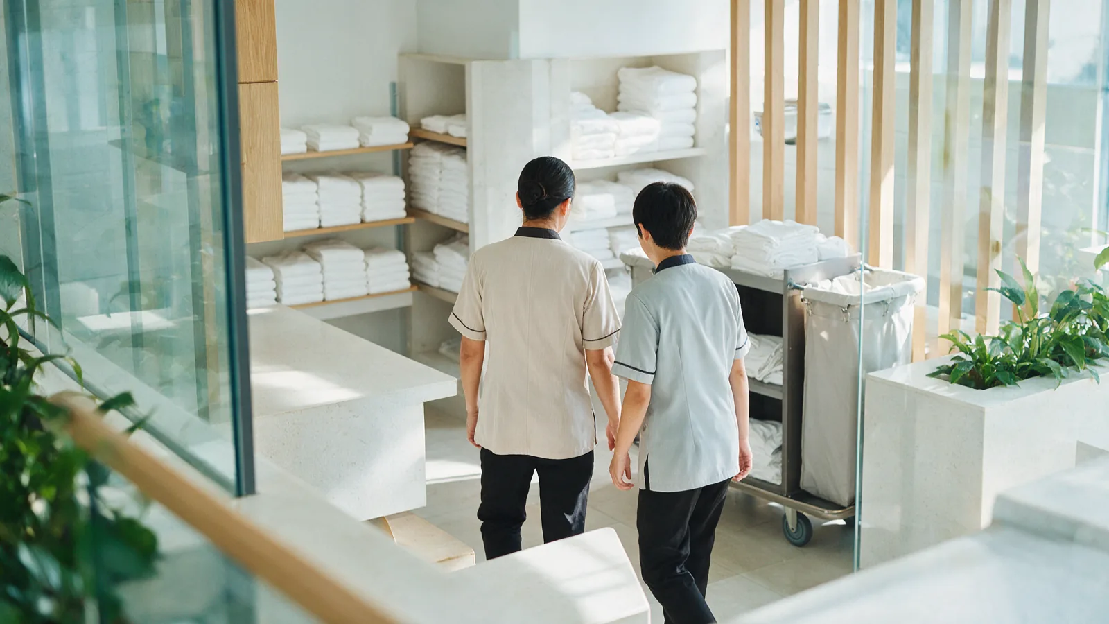 A supervisor walking a trainee in a plain uniform through a bright hotel linen room lined with folded white towels
